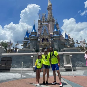 A joyful family dressed in vibrant lime green 'Dream Come True' t-shirts stands in front of the iconic Cinderella Castle at Disney World. The girl, seated in a wheelchair, is flanked by her smiling parents on a clear sunny day, with fluffy white clouds adorning the blue sky above.