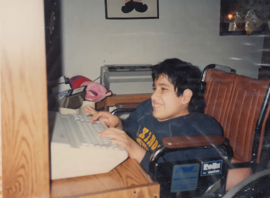 A boy from 1987 grins broadly as he sits at his new computer system and desk, a fulfillment of his wish by Dream Come True, with vintage technology and décor surrounding him.