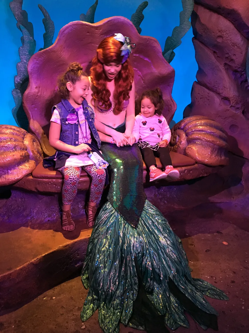 User Two young girls and a mermaid sitting in a themed setting that resembles an underwater scene, with the mermaid character engaging happily with the children.